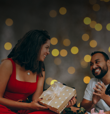 Woman in a red dress holding a gift box with a man in a white shirt, surrounded by warm bokeh lights.